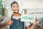 Portrait of one young hispanic man showing thumbs up while holding a "hiring" sign at a window on display in a cafe or store. Mixed race guy advertising job opportunity while recruiting new staff for his startup shop