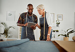 Two clothing designers using a digital tablet while working with material at work. Young african american tailor talking and holding a digital tablet while working with a female caucasian colleague