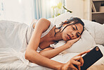 A young mixed race woman looking depressed while scrolling social media and lying in bed. An attractive Hispanic female using her cellphone and looking exhausted while resting in her bedroom