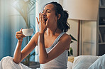 A young mixed race woman yawning while lying in bed and drinking coffee. An attractive Hispanic female waking up from a her sleep and getting ready to begin her day