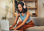 Beautiful mixed race woman using laptop for blogging while listening to music on headphones in living room at home. Hispanic entrepreneur sitting alone on lounge sofa, typing blog post on technology