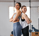 Portrait of two confident young women standing back to back with arms crossed while exercising in a gym. Determined female athletes and friends looking happy and motivated for training workout together