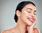 A beautiful smiling happy mixed race woman applying cream to her face. Hispanic model with glowing skin against a grey copyspace backgroun