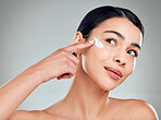 A beautiful smiling happy mixed race woman applying cream to her face. Hispanic model with glowing skin against a grey copyspace background