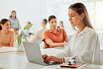 One young hispanic business woman working on a laptop in a busy office with her colleagues in the background. Focused entrepreneur browsing the internet while planning ideas at her desk in a creative startup agency