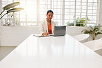 Young mixed race businesswoman writing in a notebook while working on a laptop at a table in an office at work. Serious hispanic businessperson taking a note in a diary using a laptop while working in corporate