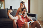 Funny romantic couple pulling faces and fooling around while holding a mobile phone and taking a selfie together. Playful man and woman sitting together and taking picture or doing video call together