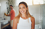 Young caucasian couple sharing a bathroom with focus on happy young woman smiling while looking at the camera in the foreground while waiting to get ready. Boyfriend washing hands in background