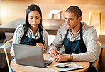 Two young mixed race employees wearing aprons sitting at a table in a coffee shop using a laptop and calculator at work. Young male and female hispanic colleagues brainstorming as a team because alone we can do so little, together we can achieve so much