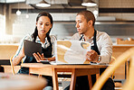 Two restaurant workers planning on a digital tablet in a small business. Young mixed race colleagues reviewing plans and reports in a cafe. Colleagues doing digital stocktake and orders with suppliers