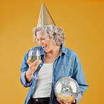 One happy mature caucasian woman holding a disco ball and drinking a glass of white wine while wearing a birthday hat against a yellow background in the studio. Smiling white lady celebrating