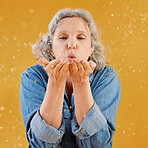 One happy mature caucasian woman blowing confetti out of the palm of her hands while posing against a yellow background in the studio. Smiling white lady showing joy and happiness while celebrating