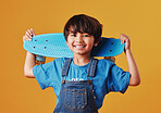 An adorable little asian boy looking happy while holding his skateboard against an orange background. Cute boy wearing casual clothes smiling as he carries his skateboard. Cute child having fun