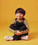 A cute little asian boy sitting on the floor with casual clothes while laughing and crossing his legs against an orange copyspace  background. Adorable happy little boy safe and alone