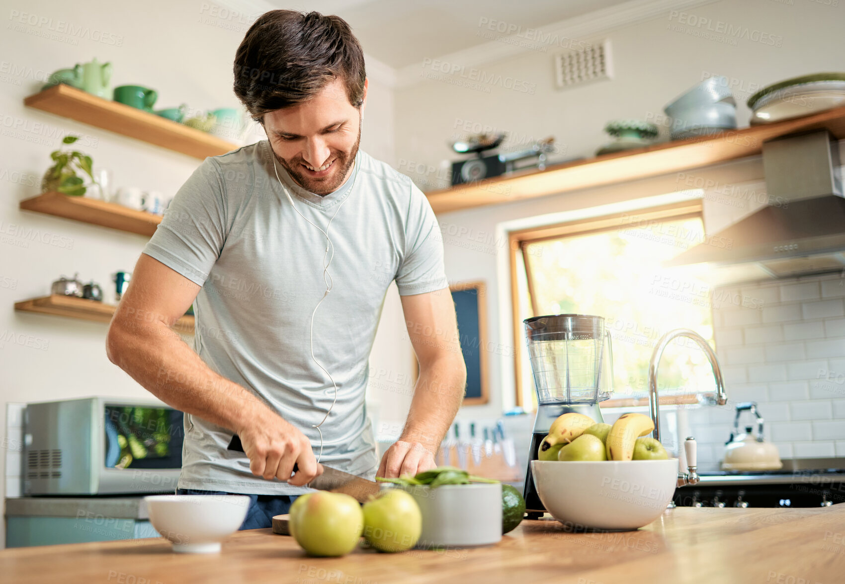 Buy stock photo One fit young caucasian man cutting ingredients with knife to make healthy green detox smoothie while wearing earphones in kitchen at home. Guy having fresh fruit juice to cleanse and provide energy for training. Wholesome drink with vitamins and nutrients