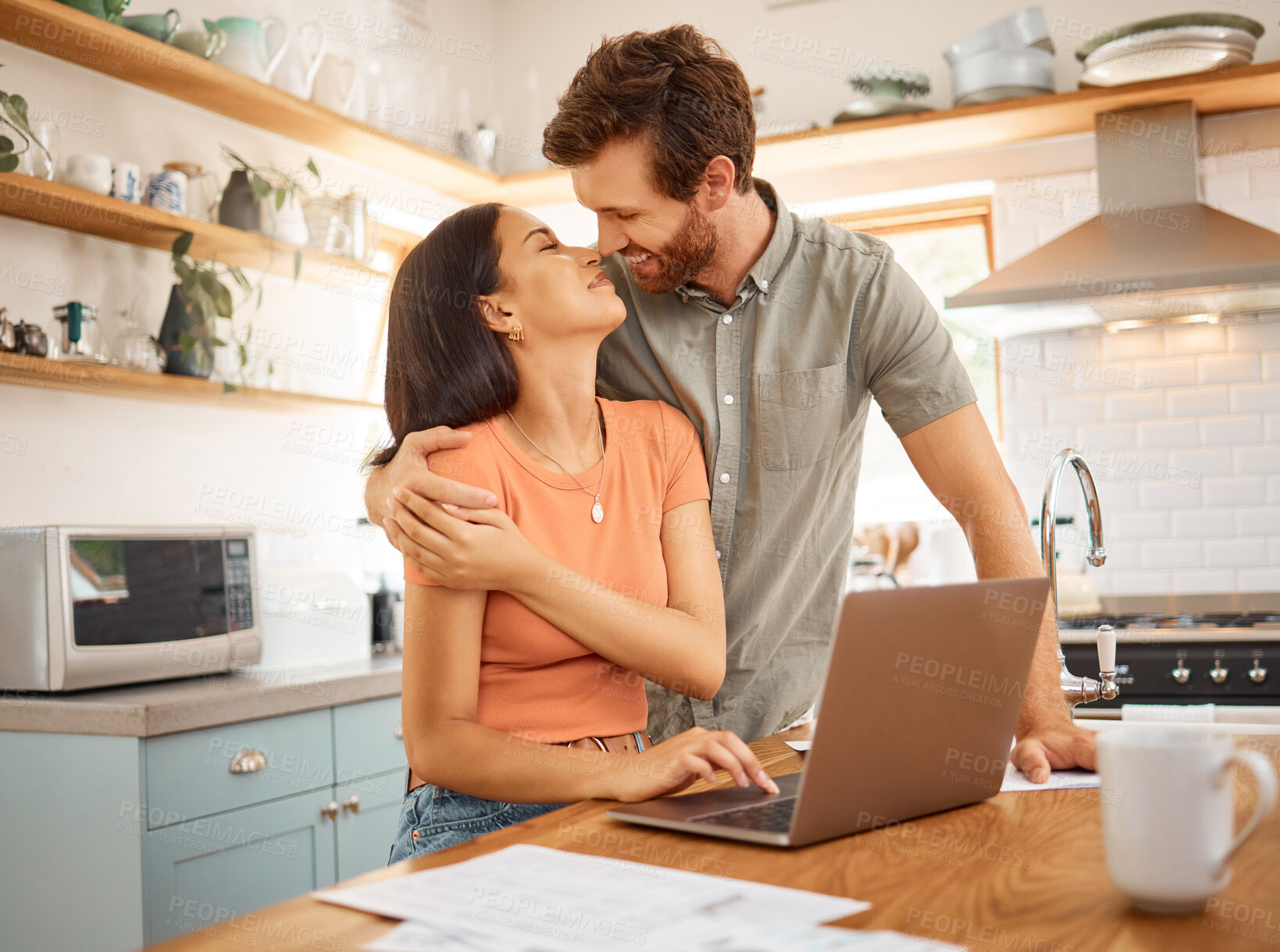 Buy stock photo Young happy interracial couple bonding while going through paper and using a laptop together at home. Caucasian boyfriend and girlfriend hugging in the kitchen. Cheerful husband and wife working on a laptop together