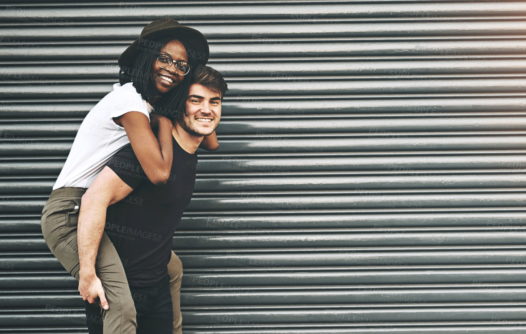 Buy stock photo Fun, trendy and happy interracial couple enjoying a piggyback ride together outside against an urban background. Lovers bonding and being playful. Smiling and relaxing while walking outdoors