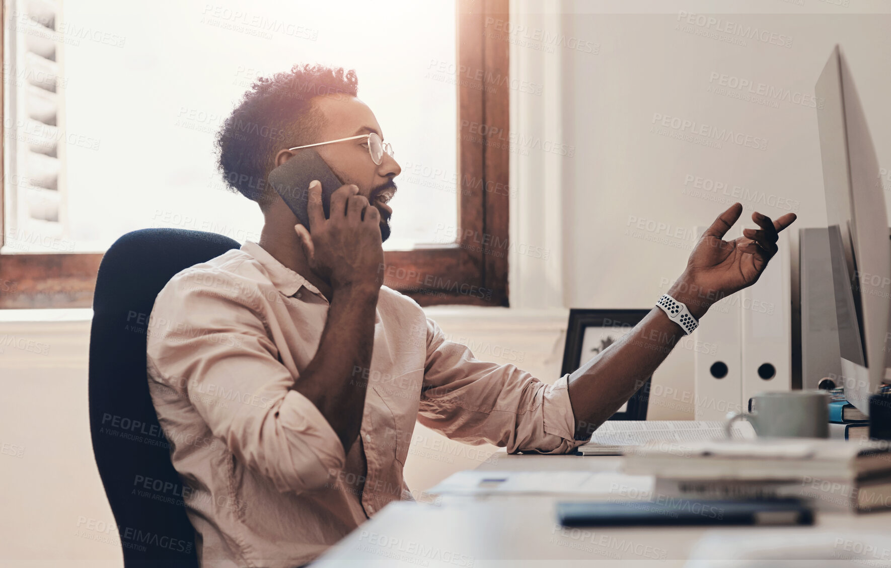 Buy stock photo Shot of a young businessman talking on a cellphone while working on a computer in an office