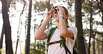 Woman, backpack and hiking with binoculars in forest for bird watching, sightseeing or outdoor discovery in nature. Female person, tourist or hiker with vision for tourism, trekking or observation