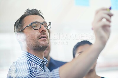 Buy stock photo Cropped shot of a handsome businessman making notes on a glass wall while his colleagues look on