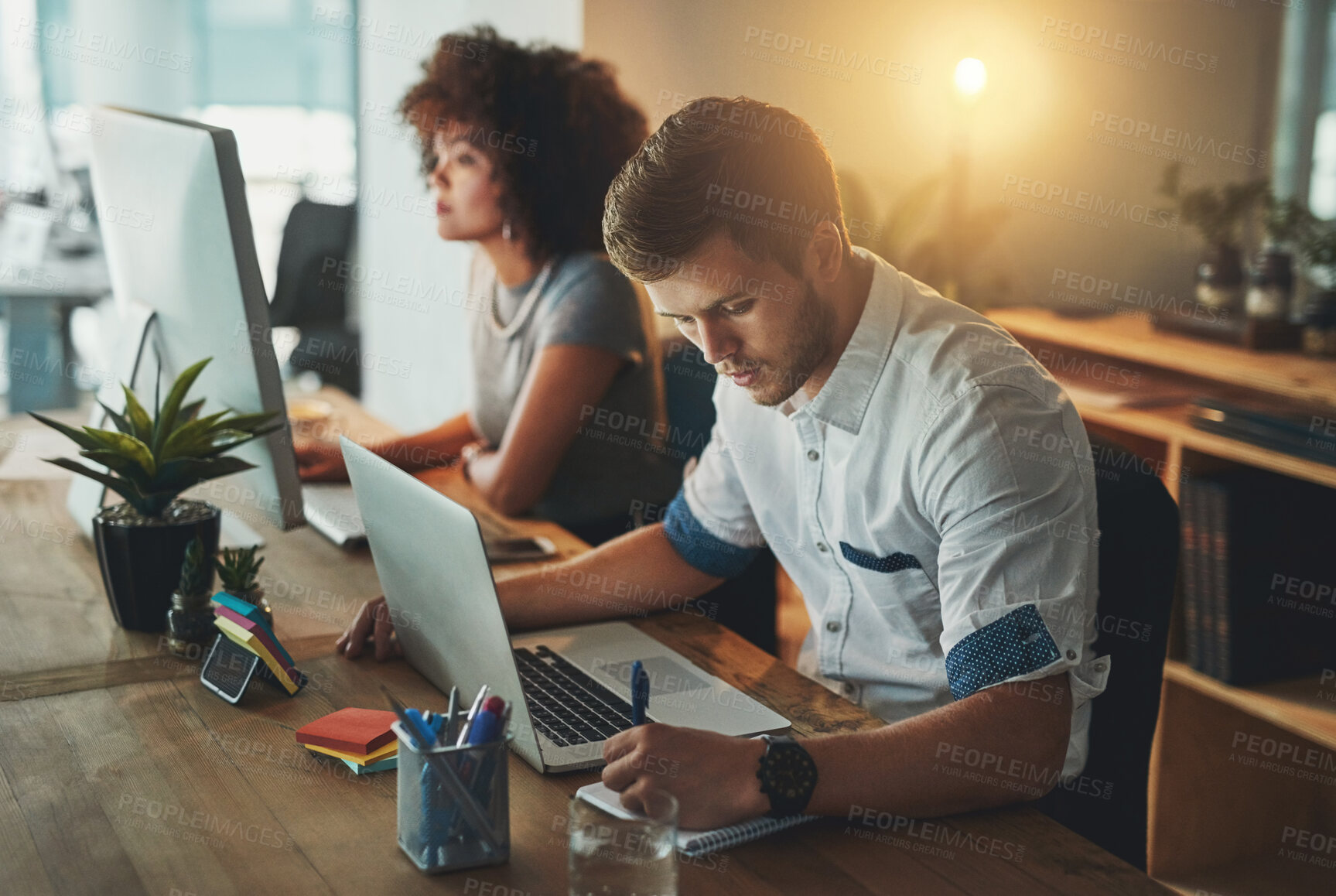 Buy stock photo Shot of a young designer working late with his colleague blurred in the background