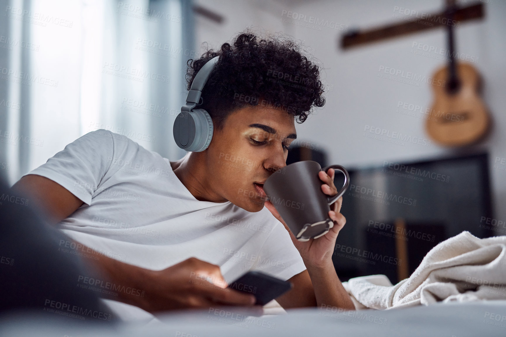 Buy stock photo Drinking coffee, phone and man with headphones for music, morning or scroll on social media to relax. Mobile, tea and person in bed for streaming sound, text message or listen to rock in bedroom home