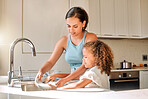 Little girl helping her mother with household chores at home. Happy mom and daughter washing dishes in the kitchen together. Kid learning to be responsible by doing tasks