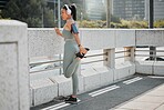 Young mixed race hispanic female athlete wearing headphones and listening to music while stretching her leg before a run outside in the city. Stretching is important to prevent injury