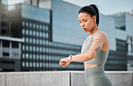 Young hispanic fit female athlete with tattoos looking serious while checking the time on her watch to track her progress while taking a break from working out in the city. Exercise is good for your health and wellbeing