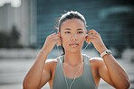 Young serious looking mixed race female athlete wearing earphones and listening to music while standing outside in the city. Positive, focused, workout. Taking a break