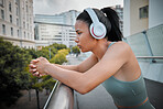 Young serious mixed race hispanic female athlete looking thoughtful wearing headphones and listening to music while standing on a bridge outside In the city. Taking a break from her excising. Determined to achieve my goal