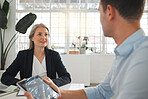 Two businesspeople talking in a meeting together at work. Business professionals talking and planning in an office. Mature caucasian businesswoman and young businessman sitting and talking at a table