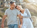 Happy and loving mature caucasian couple enjoying a romantic walk at the beach together on a sunny day. Cheerful affectionate wife resting on husband's shoulder while holding hands and bonding on vacation outdoors