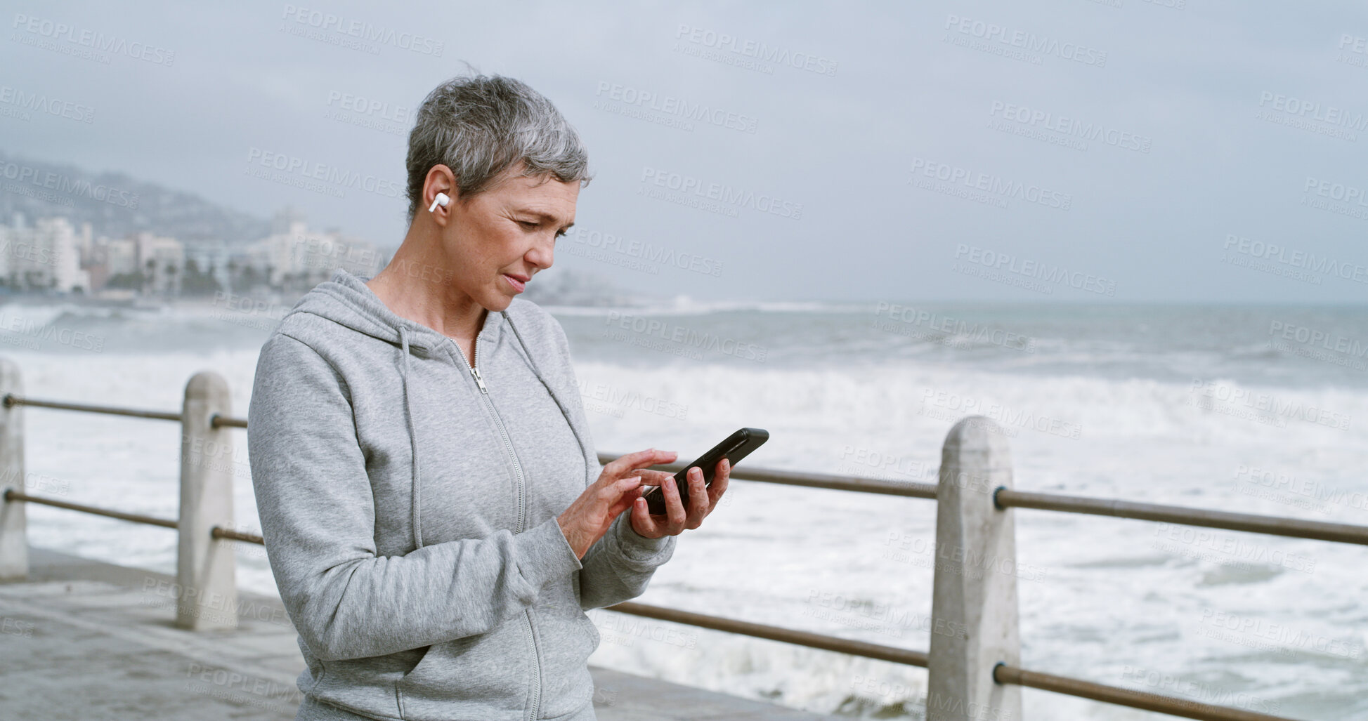 Buy stock photo Mature woman, phone and texting on promenade with earphones, reading and workout in morning. Person, smartphone and click on mobile app with running, streaming and subscription by ocean in Italy