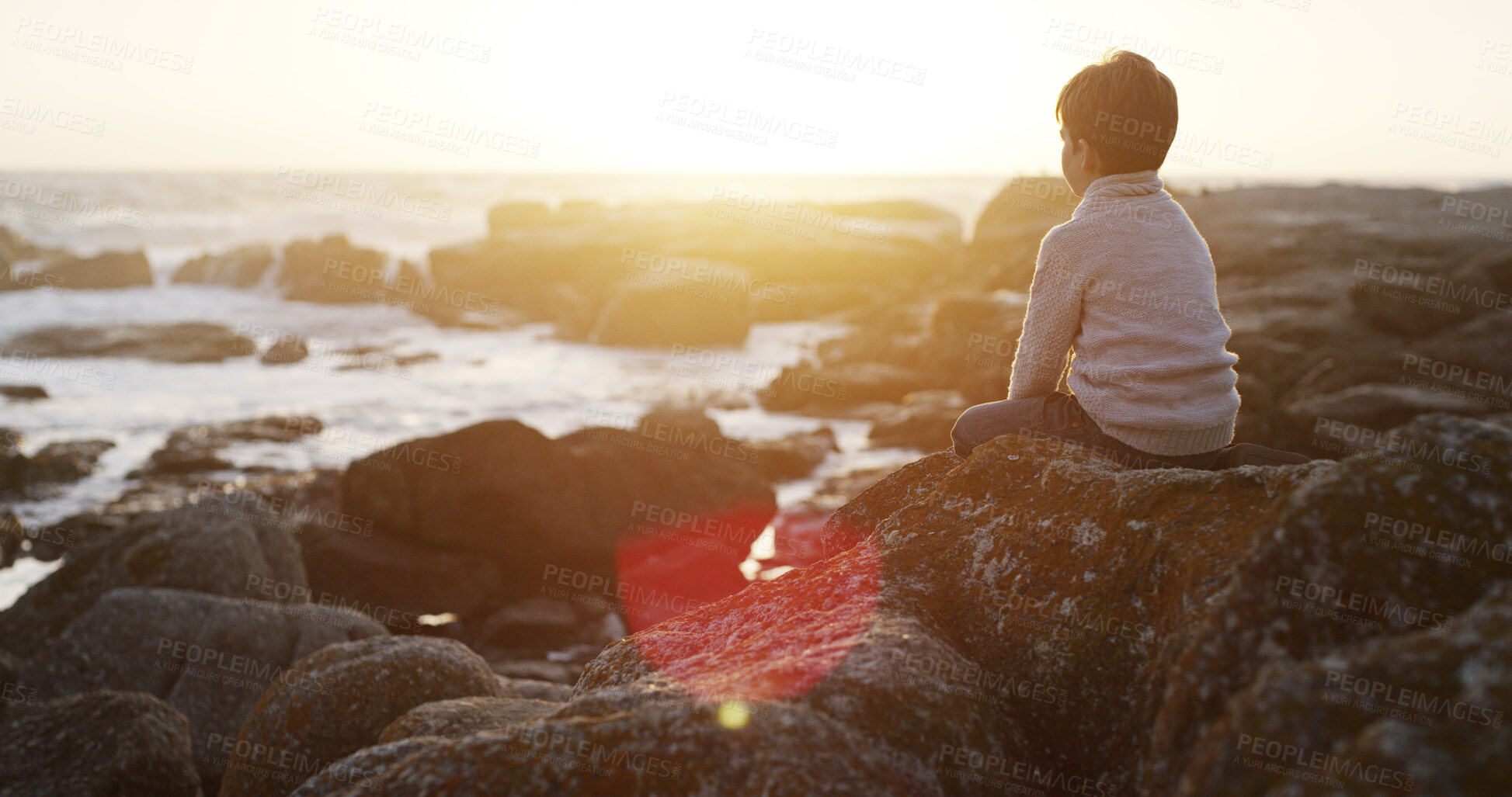 Buy stock photo Boy, beach and relax with sunset on rock for holiday, weekend or peaceful view in nature. Back view, kid or child with sunshine, waves or water for natural scenery or vacation by ocean coast or sea