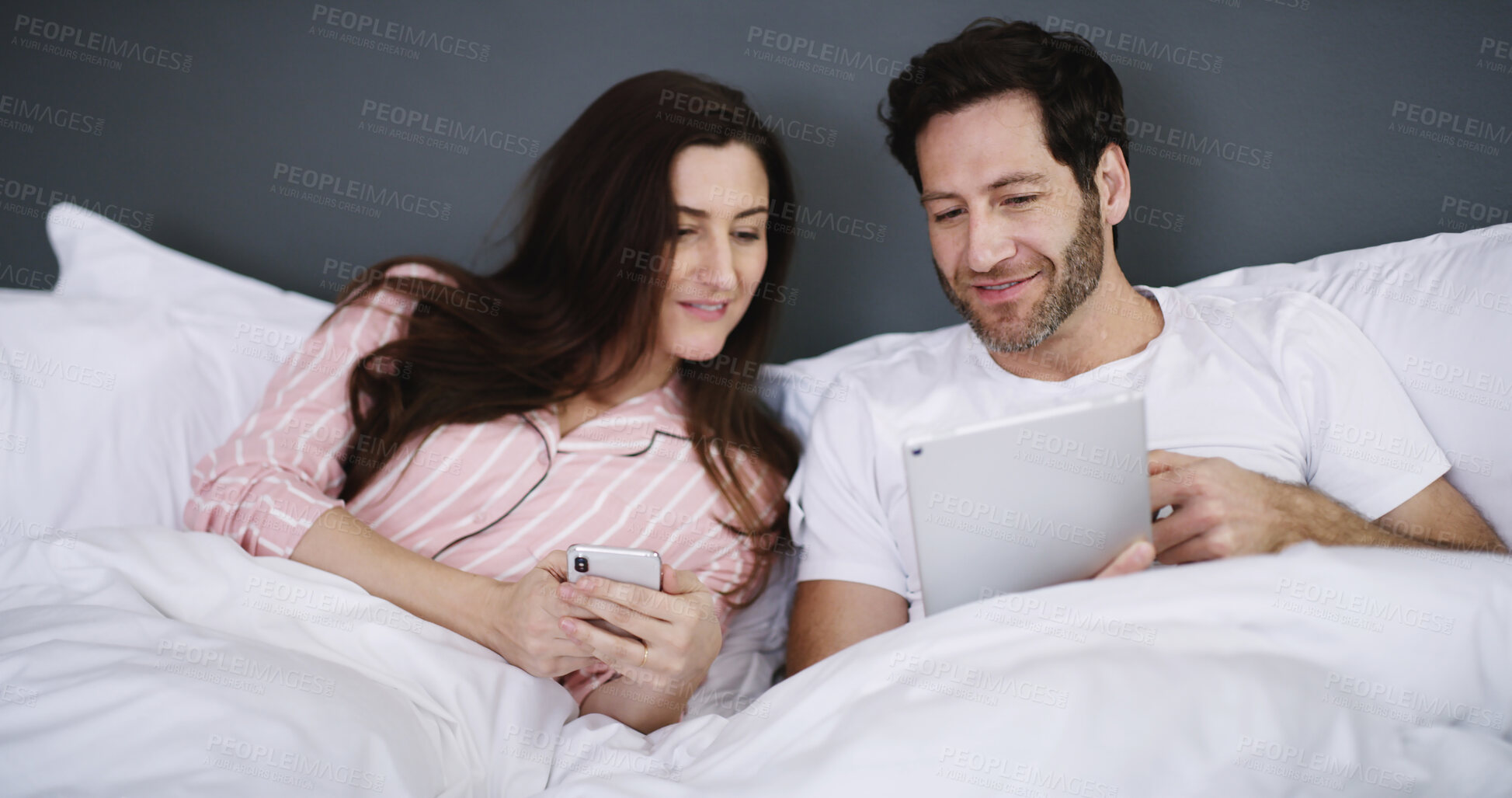 Buy stock photo Cellphone, tablet and couple resting in bed in home for networking, connectivity or browsing online. Happy, relax and young people with phone and digital technology in bedroom for contact in house.