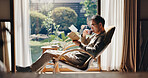 Asian man, tea and relax with book on chair for reading, story or novel by window at home. Japan, male person or reader with drink, beverage or chapter for fantasy, vocabulary or knowledge at house
