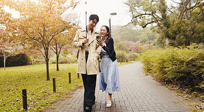 Buy stock photo Umbrella, man and woman in park with smartphone, checking schedule or online map on morning date. Phone, scroll or Japanese couple with mobile app on romantic walk in garden with cover in winter rain