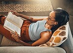 Above view of mixed race woman reading book in a living room at home. Beautiful hispanic sitting alone on lounge sofa and enjoying novel. Relaxed woman wearing glasses to read fiction story on weekend