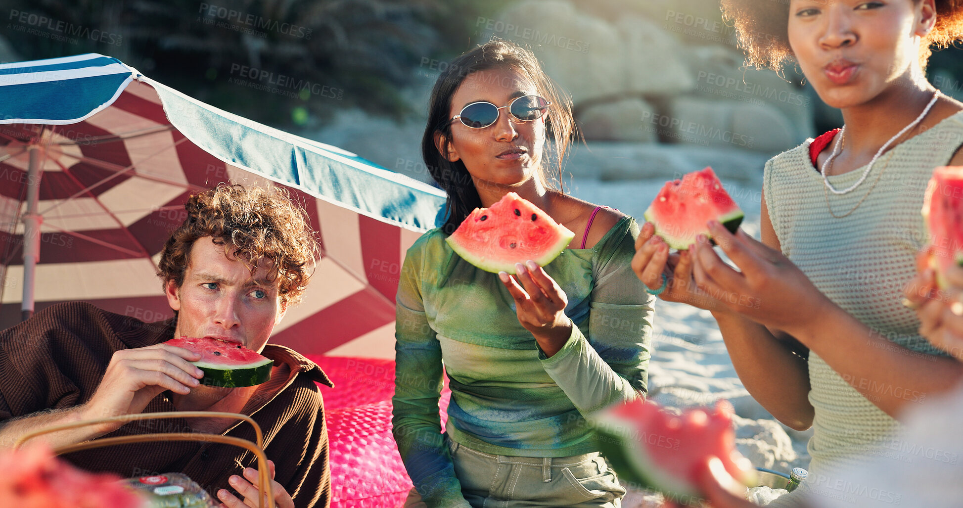 Buy stock photo People, friends and picnic with watermelon at beach, happy and conversation with reunion on vacation. Group, women and men with fruit, sharing and eating together with diversity by ocean in sunshine