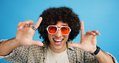 Buy stock photo Happy man, portrait or sunglasses with hairstyle or funky fashion on a blue studio background. Male person, hipster or young model with stylish shades or finger frame for summer party or festive mood