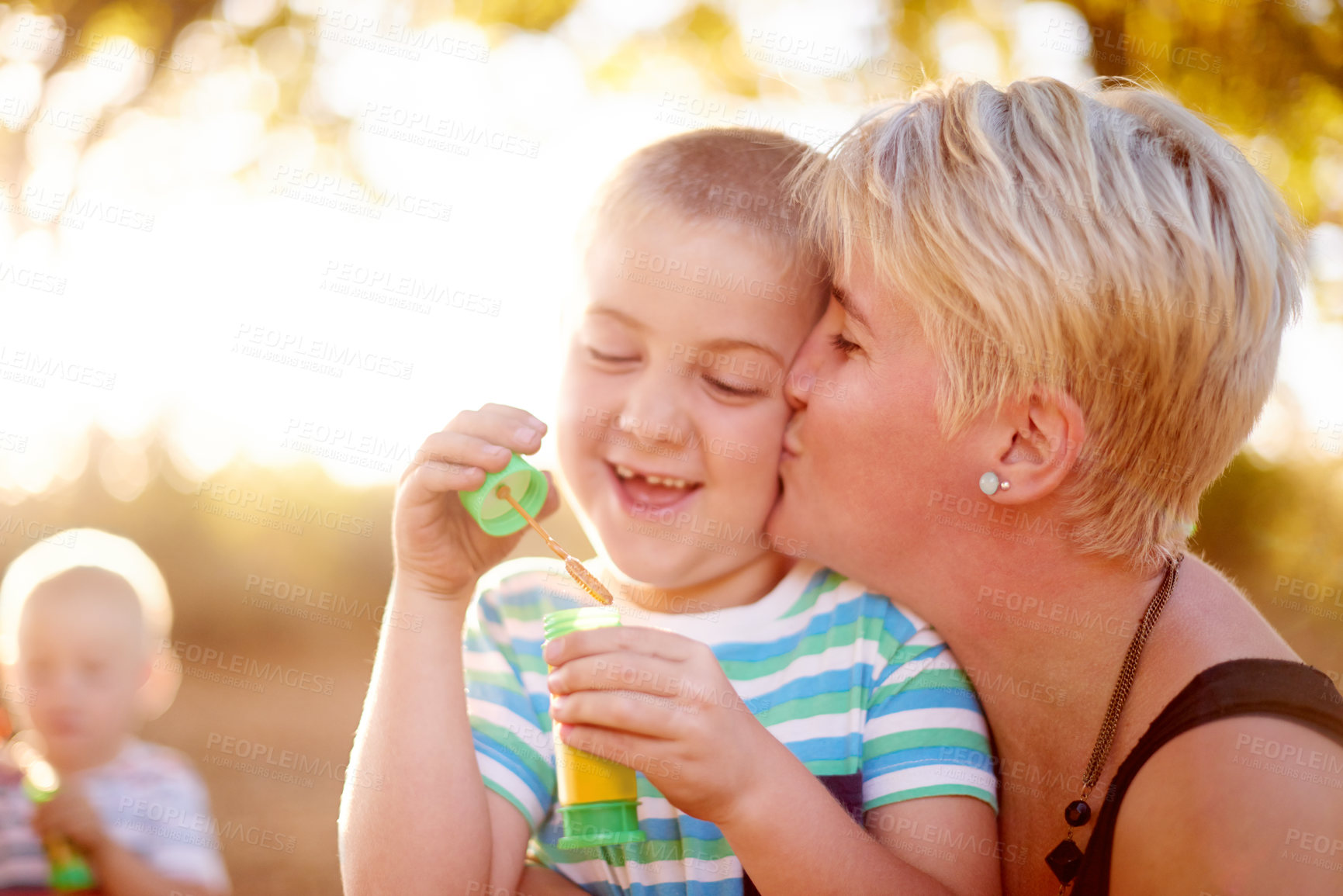 Buy stock photo Kiss, blowing bubbles and child in park with mom for fun, childhood and bonding together outdoors. Family, sunlight and woman with young boy with soap wand, toys and activity for playing in nature