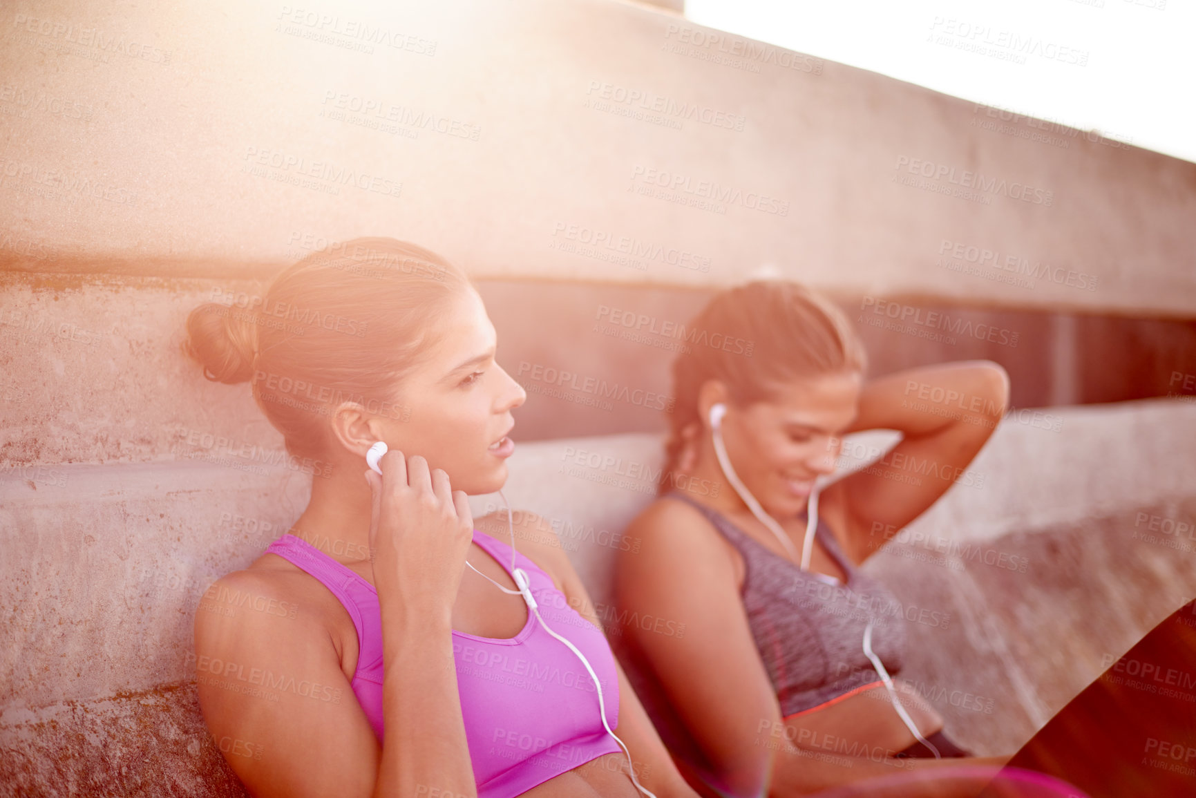 Buy stock photo Fitness, teamwork and woman with resting outdoor for cardio break, listening to music and tired of running. Twin sister, people and relax on bridge with earphones for workout radio, podcast and flare