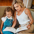 Young content caucasian mother and little daughter reading a story book together sitting on the couch at home. Carefree girl reading with her mom. Woman reading a fairytale to her child