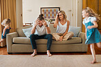 Young caucasian mother and father suffering a headache with their daughters running in the lounge at home. Little sisters being playful while their parents are stressed. Woman and man upset sitting on the couch