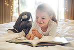 Adorable little girl reading book while lying on bed with teddybear. Cute kid reading with finger while relaxing in her bedroom