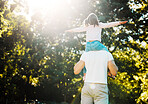 Father and playful daughter from behind having fun in sun at park with copyspace on green trees. Loving parent carrying cheerful girl on shoulders for piggyback ride with arms out pretending to fly