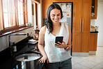 Happy mixed race woman smiling while using smart phone at home. Woman reading text message or chatting on social media while busy with chores in the kitchen. Young woman texting while spring cleaning