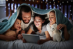 Happy caucasian family with two children using digital tablet lying under blanket in the dark at night with their faces illuminated by device screen light. Family of four reading online story or watching movie before bedtime