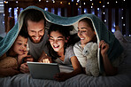 Happy caucasian family with two children using digital tablet lying under blanket in the dark at night with their faces illuminated by device screen light. Family of four reading online story or watching movie before bedtime